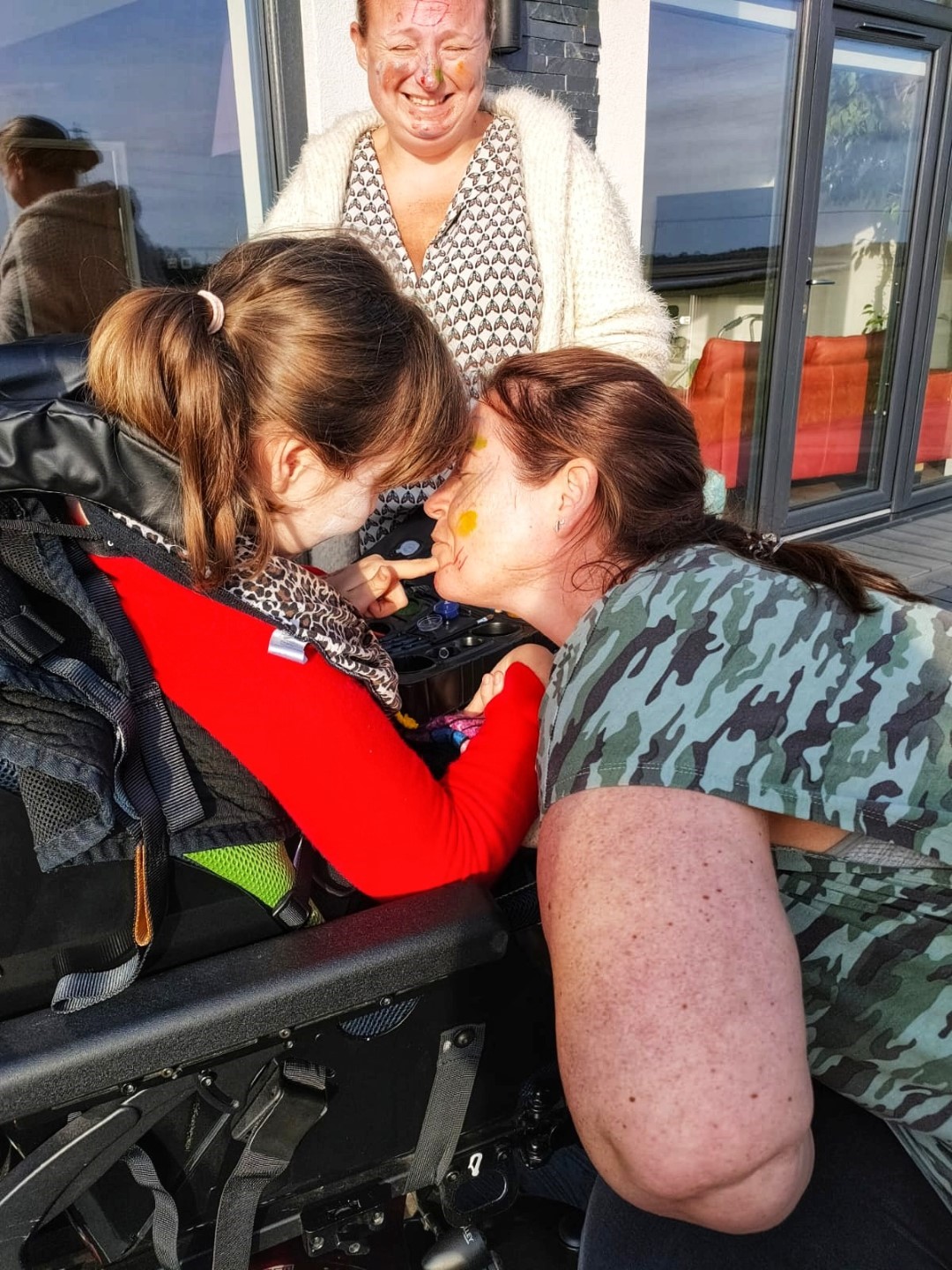 Young girl in a wheelchair touching lady's face with face paints. Lady stood to left laughing.