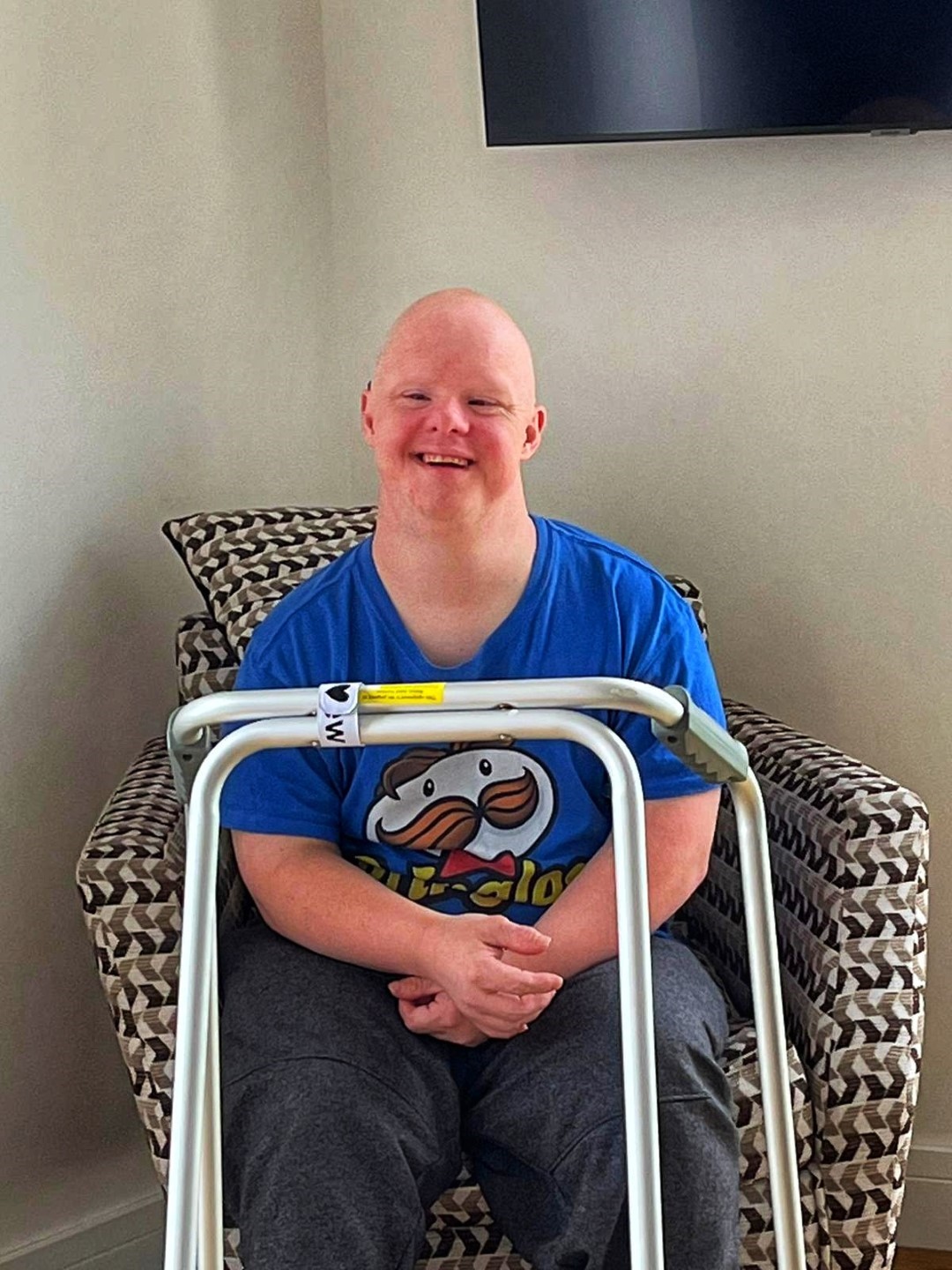A young man sat in a modern chair with a walking frame. Young man is smiling and wearing a blue t-shirt.