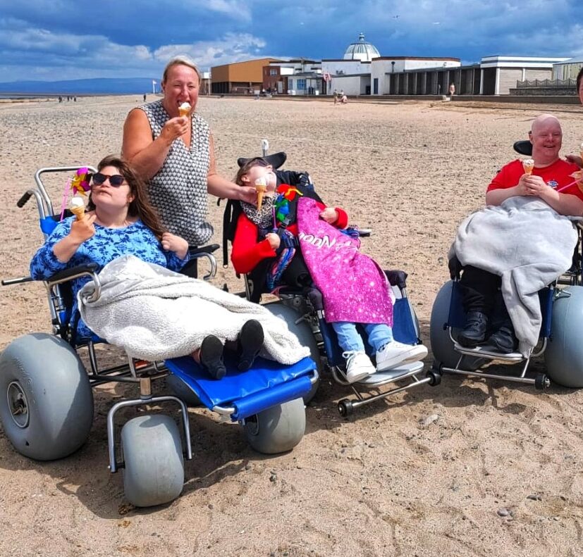 Three people in wheelchairs and one standing on a beach. Eating ice cream and enjoying themselves. Brightly coloured clothing and thrown.
