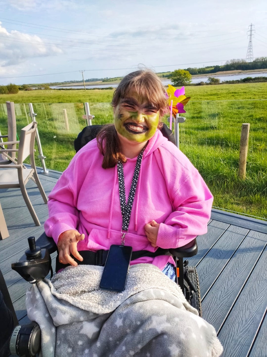 A beautiful girl in a wheelchair with green face paint and a smile that says it all. Mobile phone hanging from neck. Fields in the background and river.