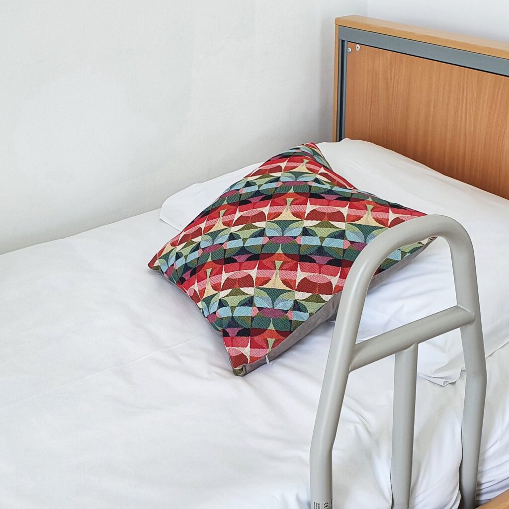 Profiling bed headboard with grab level. White bedding and brightly coloured abstract cushion.
