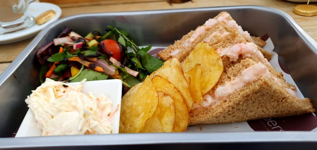 Lunch at Pipers Garstang. Prawn sandwich on brown cut in to quarters served with brightly coloured side salad, crisps and coleslaw. Served in a metal dish.