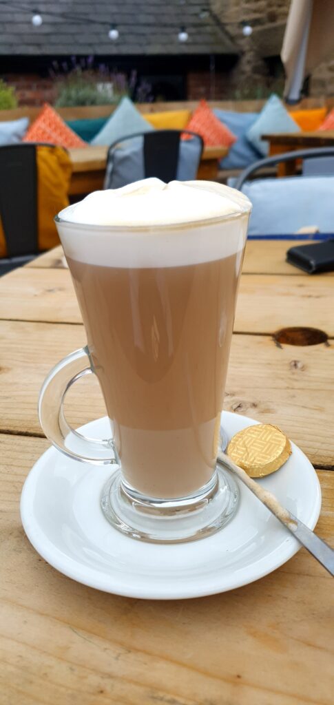 Latte served in a tall glass mug with a frothy top. Chocolate and spoon on a saucer. Served on a wooden table in the garden.
