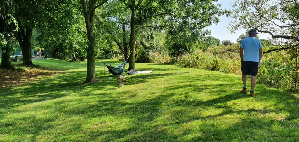Scenic Walk in Garstang Lancashire with a hammock set between two trees on the left and a person relaxing in it. Man walking on the right. Grassed area with lots of trees.