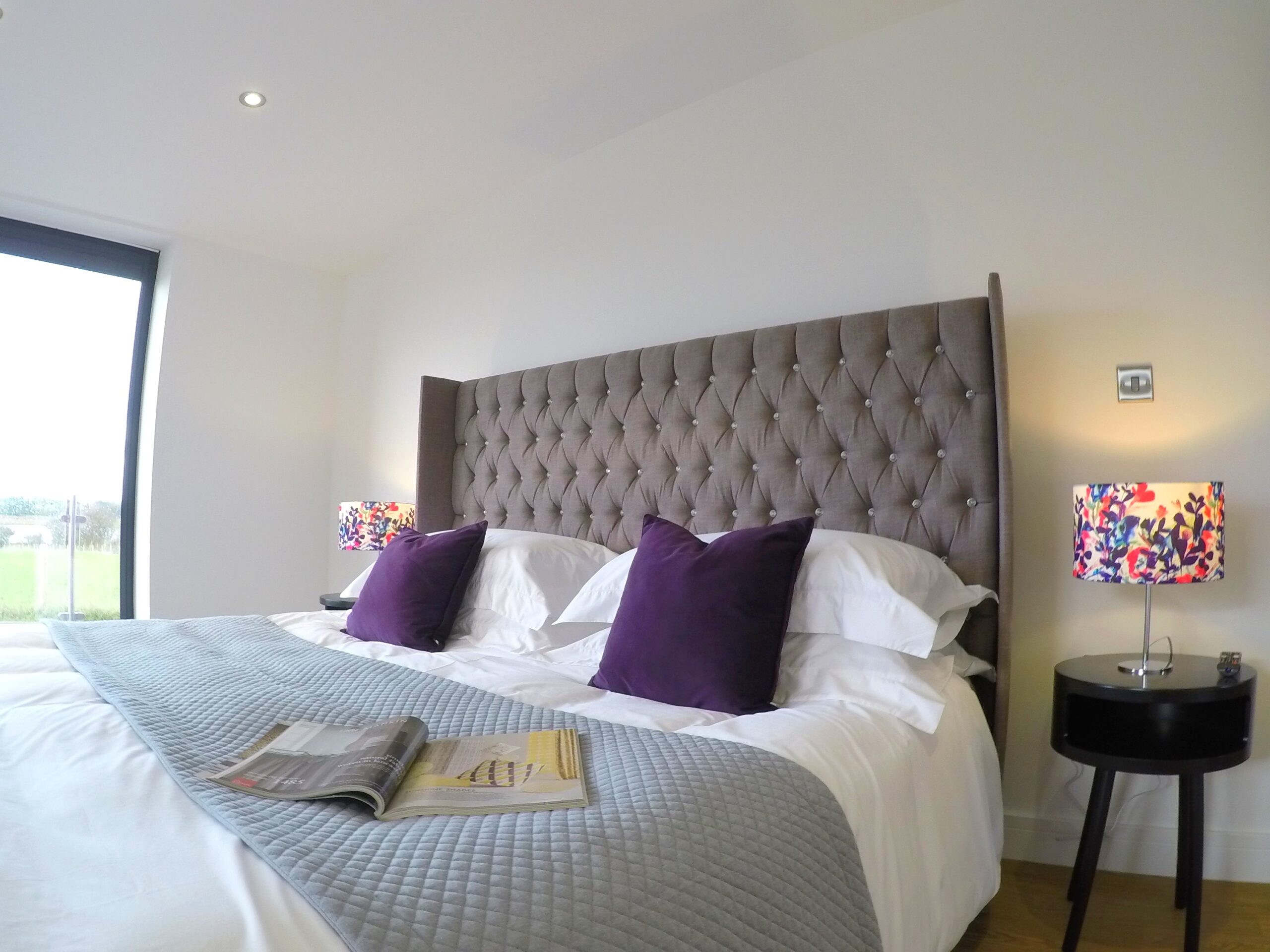 Master bedroom with misty grey headboard showing, white cotton bedding, grey throw with a magazine open. Two purple cushions. Modern black side table with open section. Table lamp with brightly coloured shade.