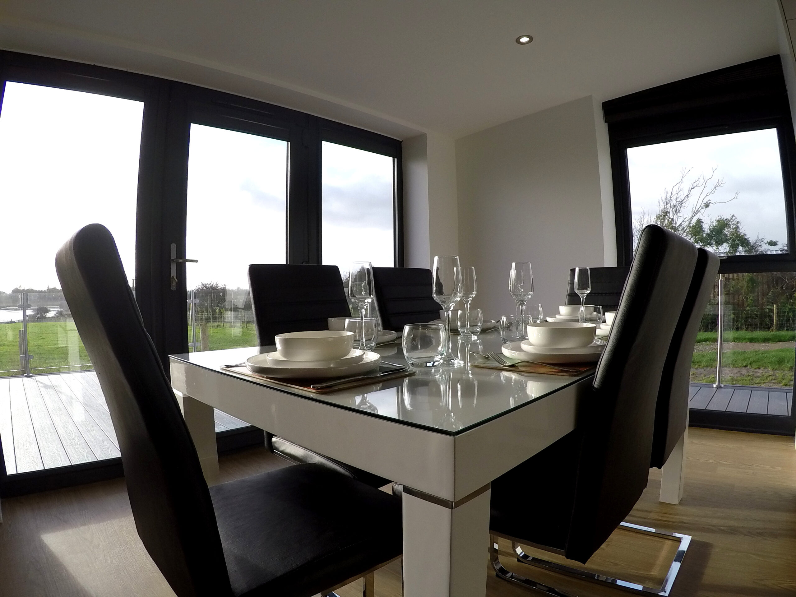 White dining table with glass top and black chairs. Table set for dinner with mats, plates and bowls and glassware. Decking views through the glass windows and door.