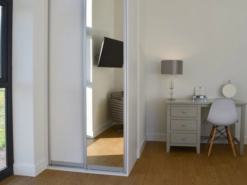Mirror reflecting modern chair in twin bedroom. Desk with three drawers with lamp with grey shade. White chair and wood effect flooring.
