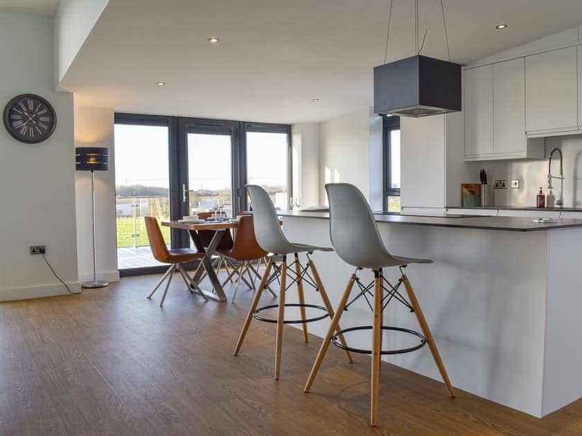 Kitchen in matt white with island. Two white bar stools. Clock to left and partial view of wood effect dining table and tan leather chairs. Wood effect flooring.