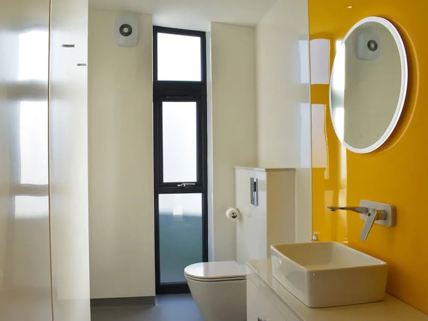 Wetroom with bright yellow wall panel. Circular mirror with light. Basin mounted on wall hung vanity with tap set into wall. Wall hung toilet. Frosted window and extractor fan.