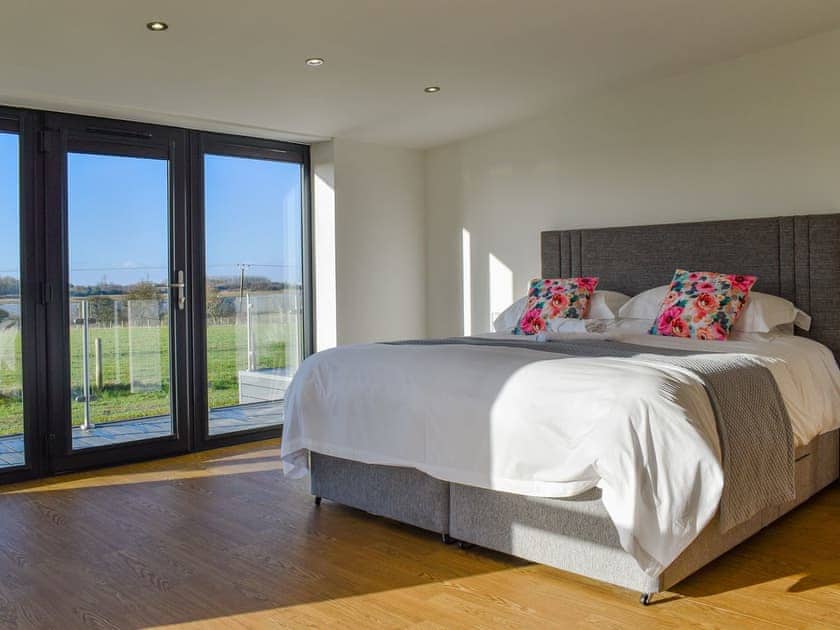 Superking bed with wood effect flooring. White cotton bedding, grey throw and bright floral cushions. Picture windows and floor leading to decking and field and river views,.