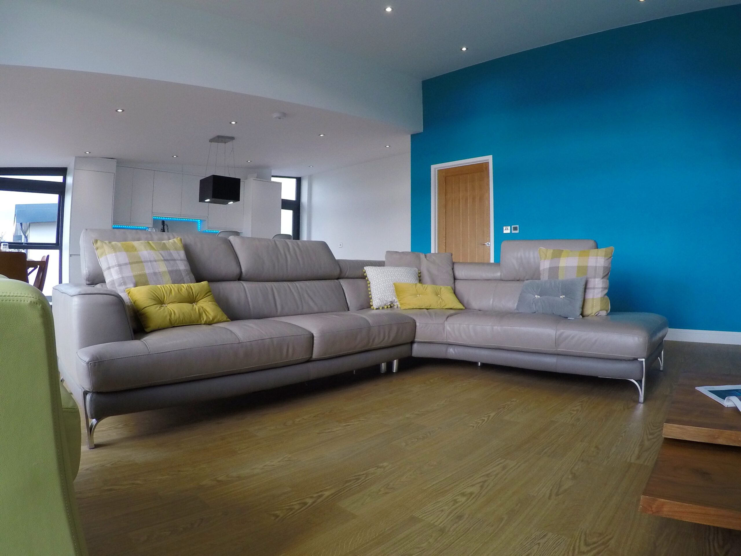 Large leather corner settee in grey with scatter cushions in check and mustard. Teale coloured wall and wood effect flooring.