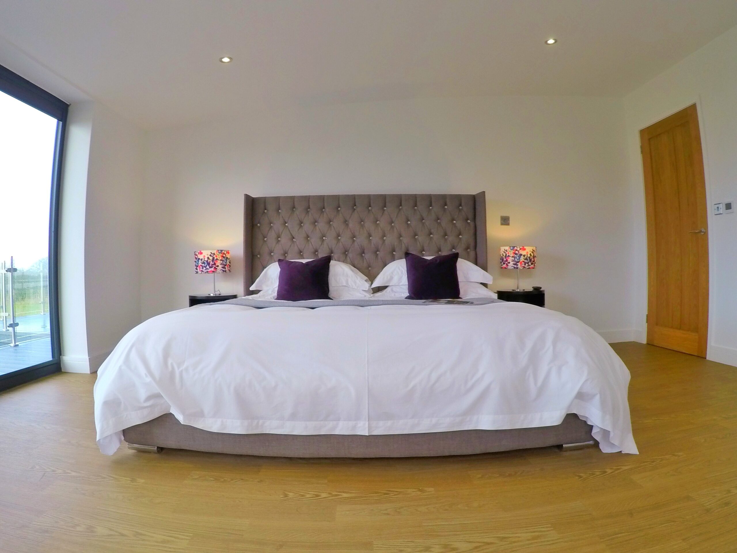 Spacious master bedroom with superking bed in misty grey with large headboard. White cotton bedding, grey throw and purple cushions. Lamps on tables next to bed with brightly coloured shades. Oak door.