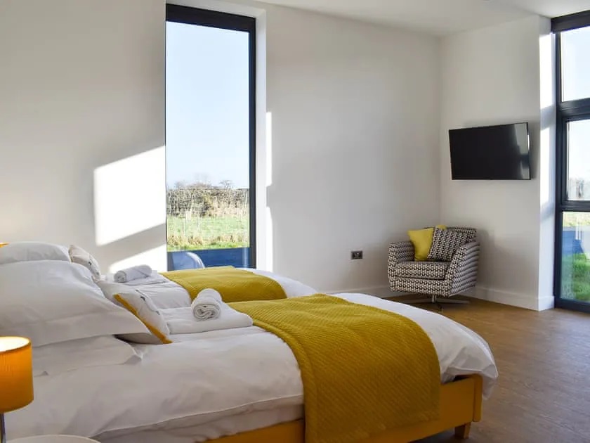 Twin bedroom with mustard coloured bed surrounds and throws. White cotton bedding. Modern chair with abstract print and yellow cushion underneath TV. Wood effect flooring. Large windows with grass views.
