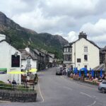Centre of Coniston with a winding road and shops and pus to either side. Silver grey car driging pass Yewdale Hotel. Blue parasols to right and yellow to the left. Coniston Old Man mountain in the background.