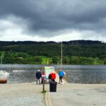 Lake Coniston close to Windermere in Cumbria with boats, a cafe and outdoor seating. Man and lady sat at a bench on the left with blue tops. A group of people and a dog walking onto the jetty, Lots of trees across the lake with a grey moody sky.