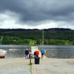 Lake Coniston close to Windermere in Cumbria with boats, a cafe and outdoor seating. Man and lady sat at a bench on the left with blue tops. A group of people and a dog walking onto the jetty, Lots of trees across the lake with a grey moody sky.
