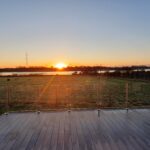 The sun setting across the river with an orange glow and shards of light. Decking with glass surround and table and chairs to the right of the decking. Field in front with sheep grazing.
