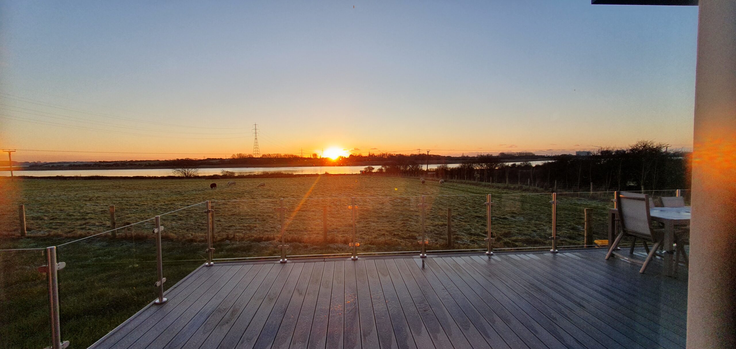 The sun setting across the river with an orange glow and shards of light. Decking with glass surround and table and chairs to the right of the decking. Field in front with sheep grazing.